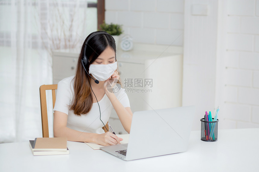 年轻女在网上戴耳机书桌上进行电子学习女孩戴耳机在家用笔记本电脑学习因特网接受新的正常教育远程和培训图片