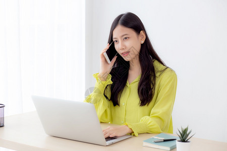 年轻的亚洲女商人在家中工作打通里的成功电话在办公桌上使用笔记本电脑的女商人在工作场所使用女智能手机的商人以及通信概念背景