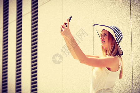 自游自在身戴太阳帽的年轻女用智能手机相拍摄自己用智能手机照相在阳光明媚的夏季日外拍摄在用智能手机拍自在外用智能手机拍自的年轻女背景