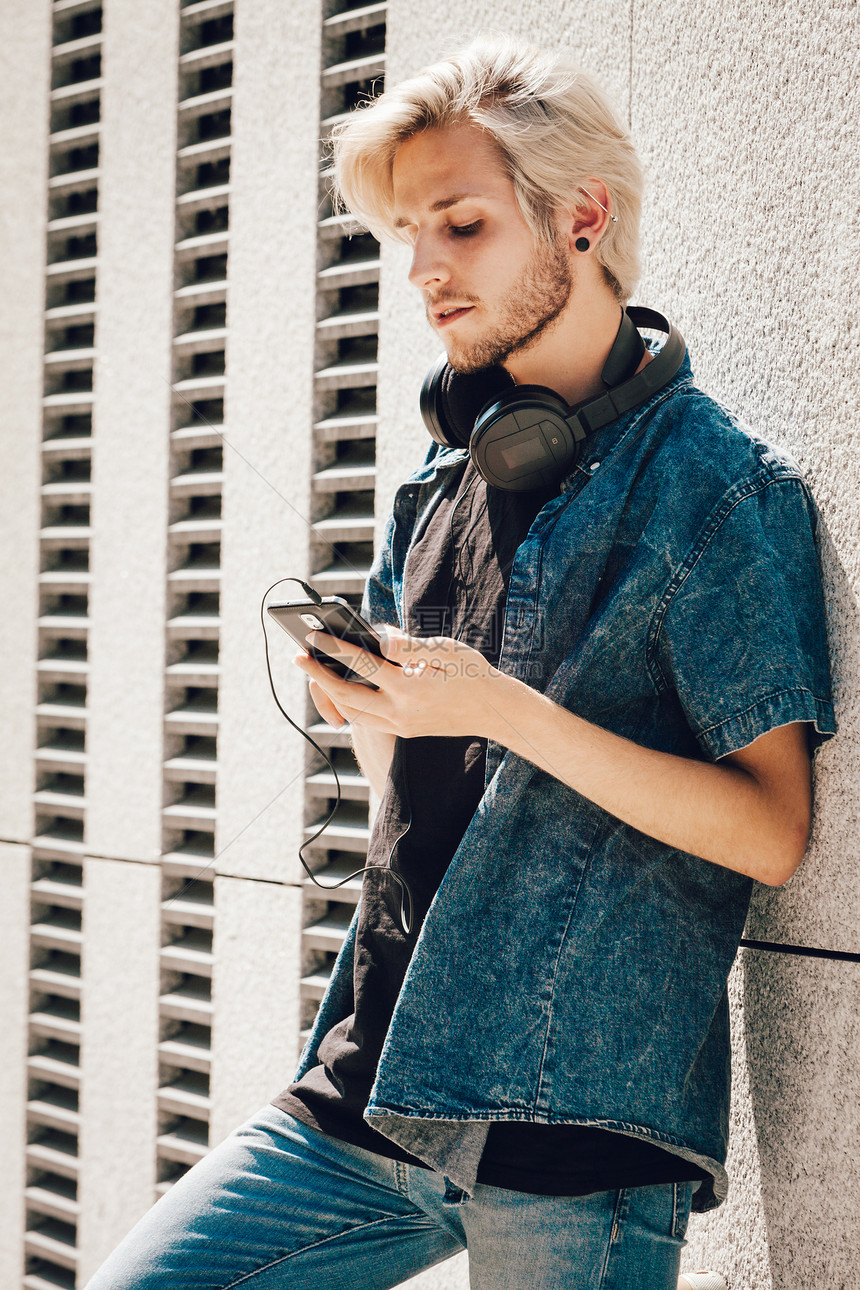 男装时尚科技都市风格的服装理念一个戴着耳机穿着牛仔裤戴着墨镜站在城市街道上看手机的时髦男人戴着耳机看手机的时髦男人图片