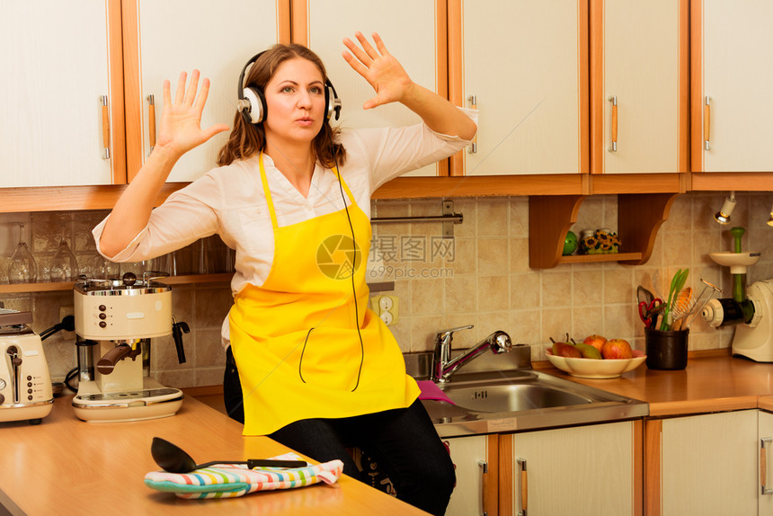在厨房里放松听音乐唱歌和跳舞快乐的家庭主妇用耳机做厨师穿黄色围裙坐在家中放松图片