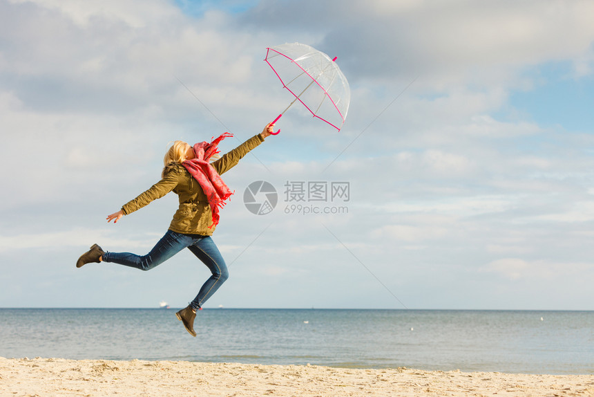 幸福享受寒冷的秋天气感受伟大的概念女人在海边滩上阳光明媚的白天和清蓝空上用透明的伞跳跃女人在海滩上用透明的伞跳跃图片