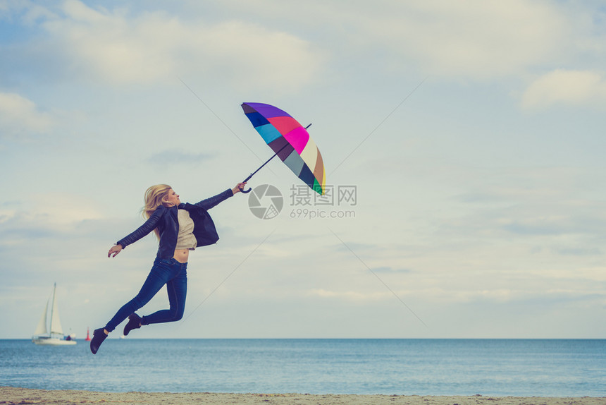 幸福享受寒冷的秋天气感受伟大的概念女人在海边滩上阳光明媚的白天和清蓝空上带着多彩的雨伞跳女人在海滩上跳图片