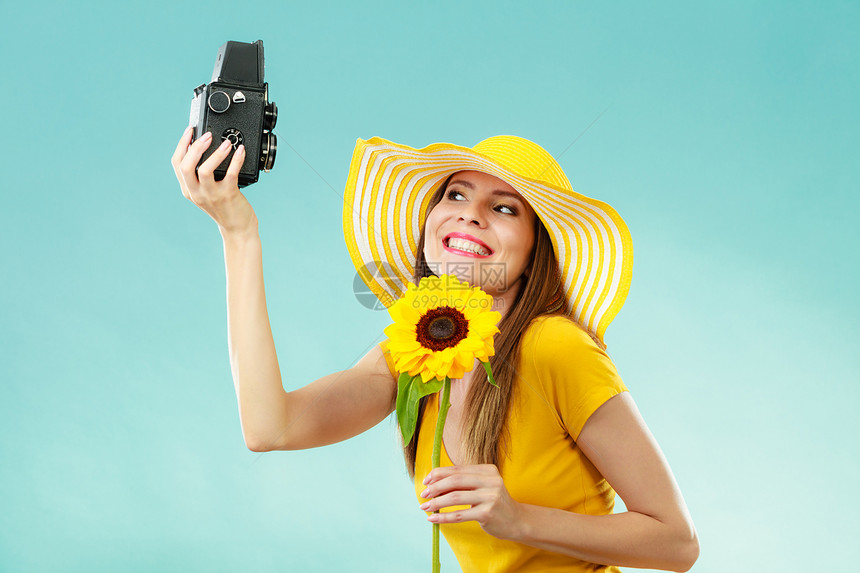 身穿黄色礼服戴向日葵帽子的暑期妇女用蓝色背景的旧相机自拍图片