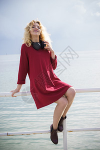 身着漂亮红裙子和戴耳机的时装女郎坐在海边的码头上脖子有耳机图片