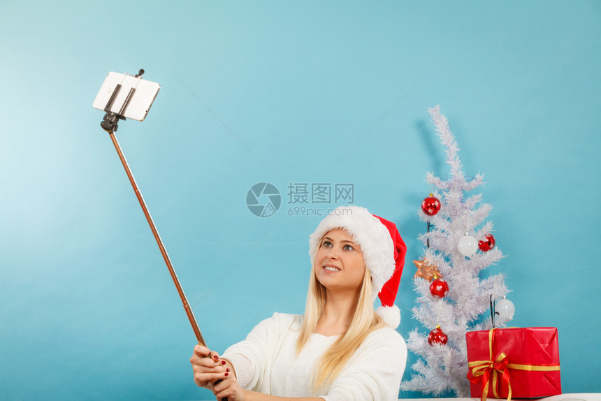 享受圣诞礼物穿着塔帽的妇女用自拍棒照己在室内拍摄蓝底照片穿着圣塔帽的妇女自拍图片
