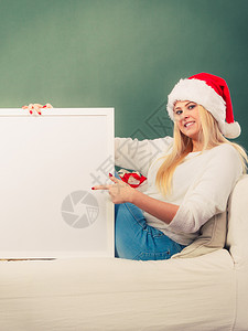 圣诞礼物和概念穿着圣诞老人帽子的金发女坐在沙上拿着白的复制空间和文字板穿着圣诞老人帽子的妇女拿着白面板背景图片