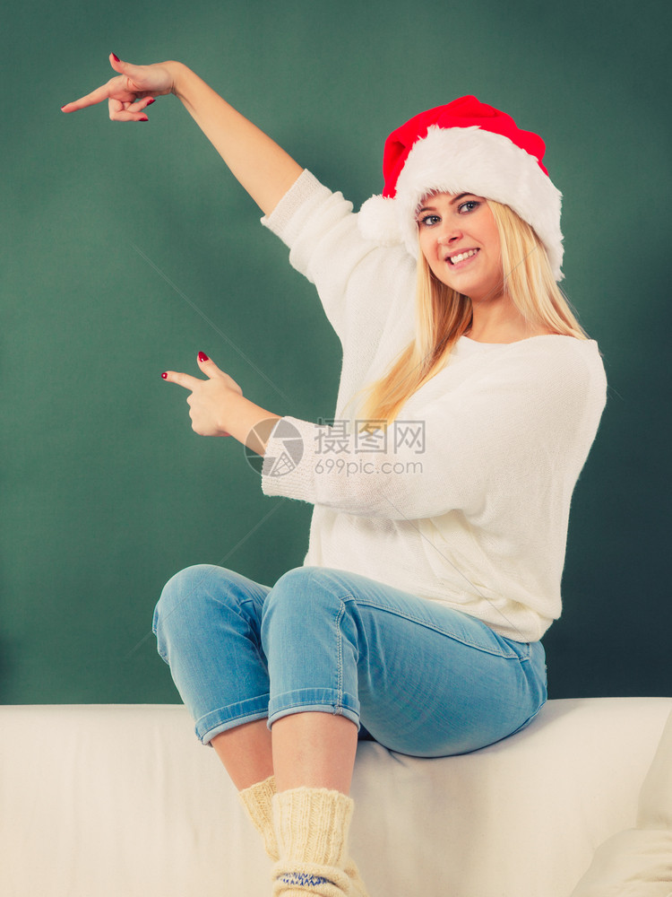 戴着圣诞帽的金发女郎坐在室内沙发上指着左边的东西戴圣诞帽的女人坐在沙发上指着什么东西图片