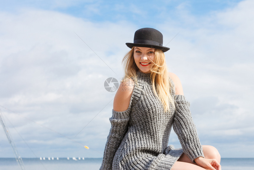 身着Fedora帽子和毛衣跳的美丽金发年轻美女假扮在天空背景上图片