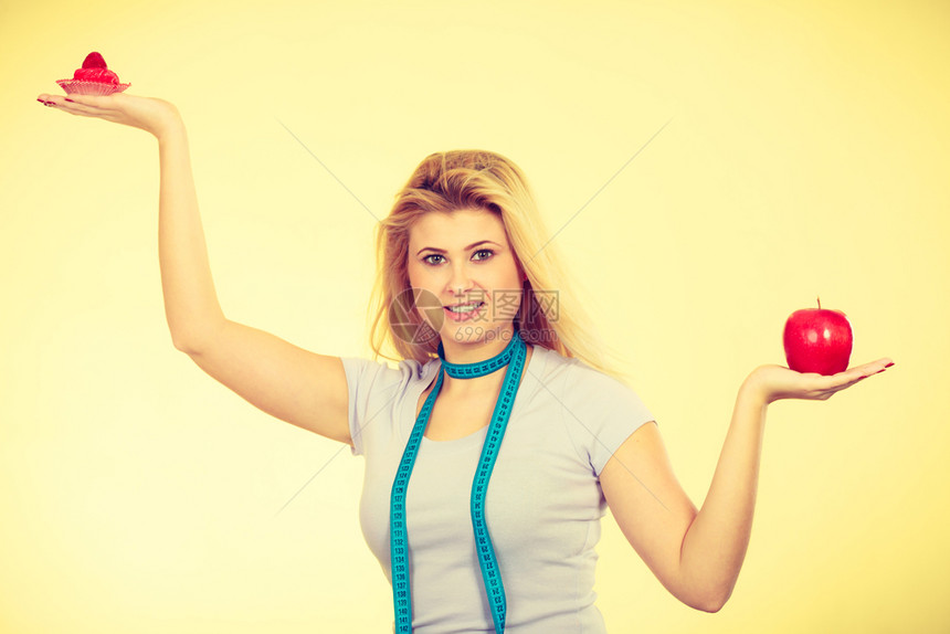 限制饮食诱惑减肥概念妇女选择甜草莓蛋糕和在脖子上贴有测量胶带的苹果工作室拍摄黄色背景妇女选择蛋糕和苹果图片