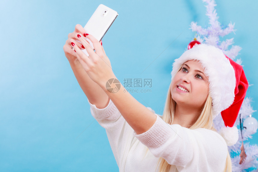 现代节日概念穿着圣诞礼帽的金发女人给自己拍圣诞照用智能手机自拍图片