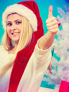 季节概念穿着圣诞帽子的金发美女做拇指手势白圣诞节树背景室内拍摄图片