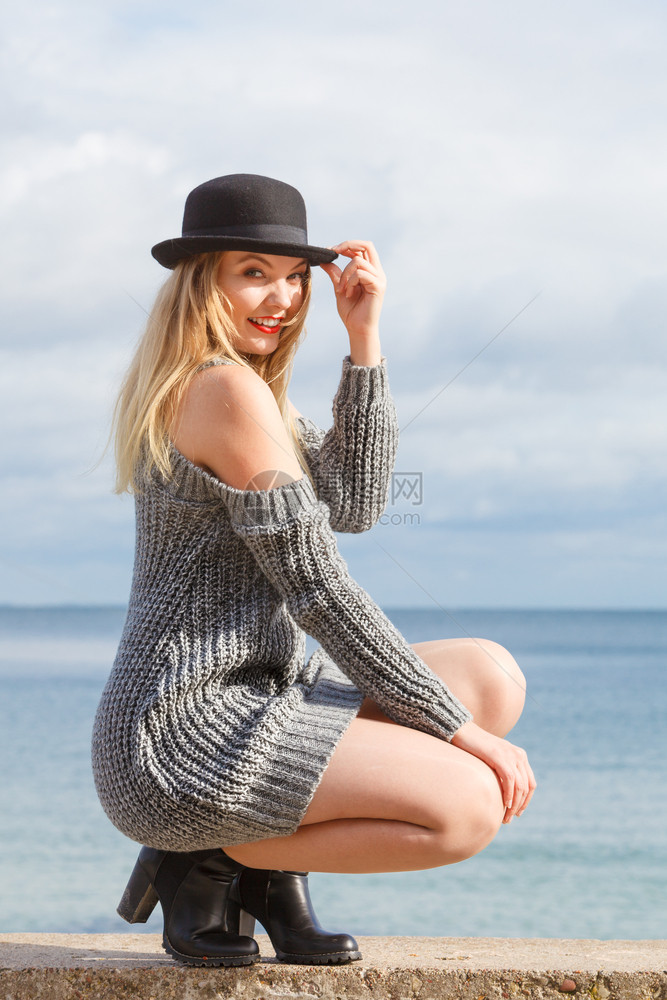 身着Fedora帽子和毛衣跳的美丽金发年轻美女假扮在天空背景上图片