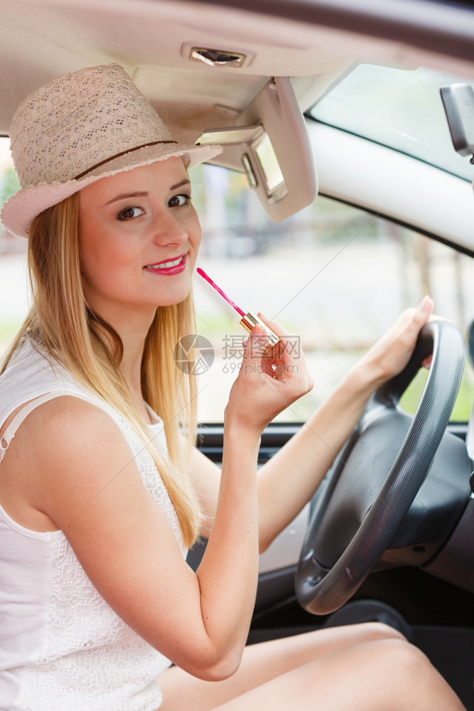 年轻有吸引力的在后视镜中照子的妇女在她嘴唇上画在驾驶汽车时涂化妆品图片