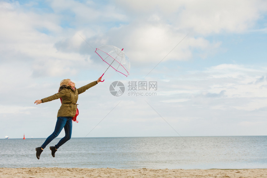 幸福享受寒冷的秋天气感受伟大的概念女人在海边滩上阳光明媚的白天和清蓝空上用透明的伞跳跃女人在海滩上用透明的伞跳跃图片