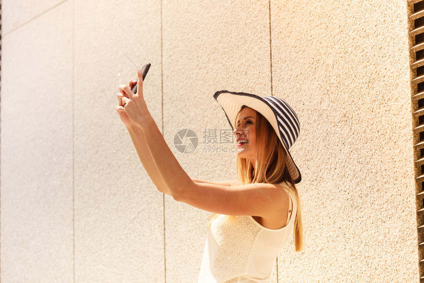 身戴太阳帽的年轻女用智能手机相拍摄自己用智能手机照相在阳光明媚的夏季日外拍摄在用智能手机拍自在外用智能手机拍自的年轻女图片