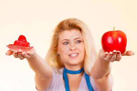 食物甜诱惑健康的选择概念妇女用测量胶带在脖子上选择苹果和甜蛋糕做决定测量胶带选择什么吃的女人背景图片