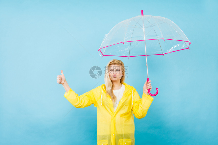 身穿黄色雨衣的金发女人拿着透明的雨伞检查天气是否雨下显示大拇指举手势图片