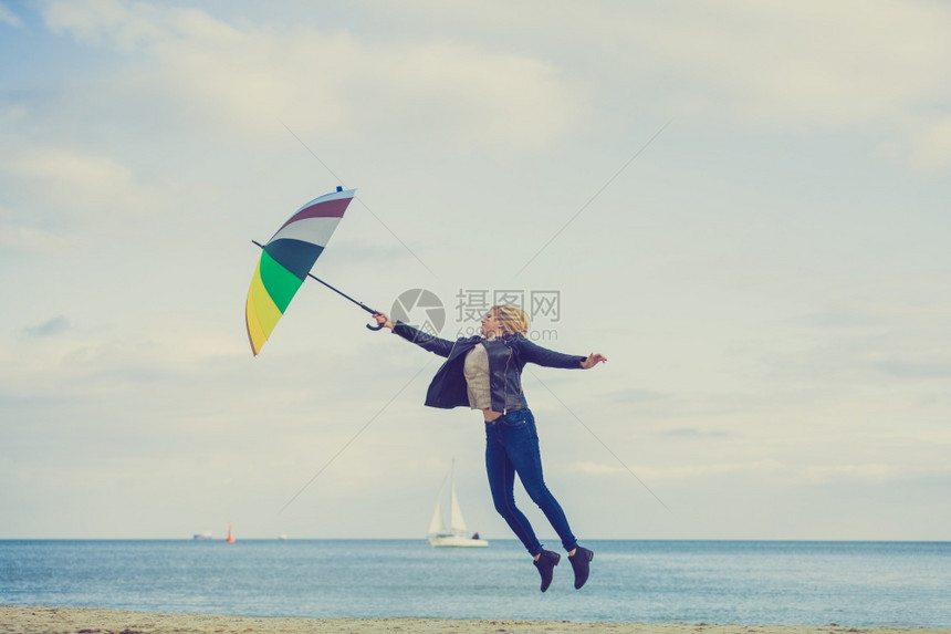 幸福享受寒冷的秋天气感受伟大的概念女人在海边滩上阳光明媚的白天和清蓝空上带着多彩的雨伞跳女人在海滩上跳图片