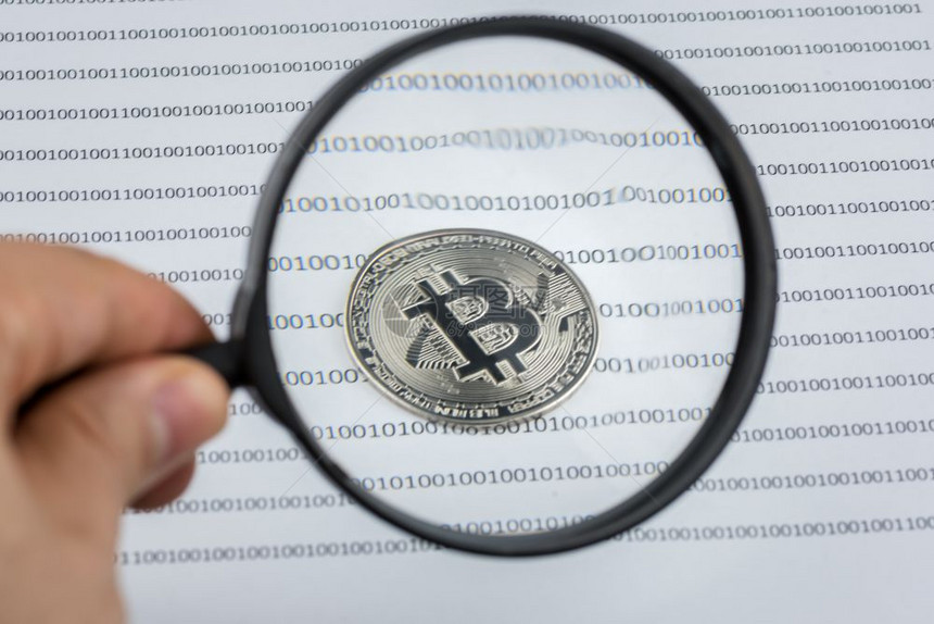 人手握着比特币的放大玻璃用零和的二元系统将纸上的硬币加密成货虚拟加密货币概念接受加密货币的图像图片