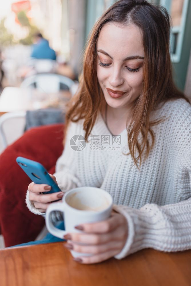 在跳跃者中美丽的漂亮年轻女孩坐在咖啡馆里使用智能手机和短信图片