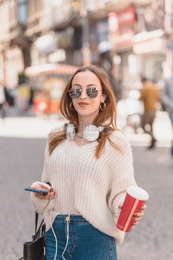 穿跳衣和牛仔裤的漂亮女孩带外送的咖啡耳机和太阳眼镜在步行时使用智能电话图片