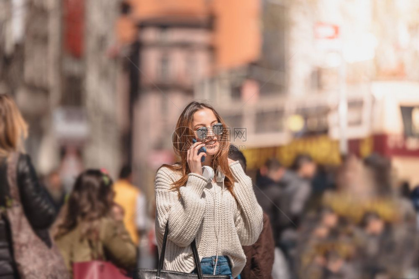 穿着带耳机和太阳眼镜的跳伞牛仔裤和的漂亮年轻女孩在人群中行走时使用智能电话图片