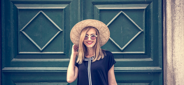 身着时尚黑衣装圆环太阳镜和黄帽子的年轻有吸引力女子在街对面墙壁上露背景图片