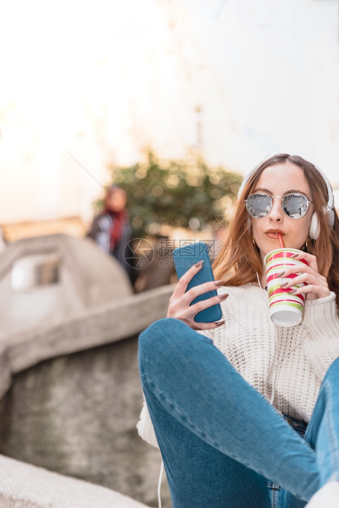 带着耳机和太阳眼镜的年轻美女的肖像在拿着果汁和坐时使用智能电话图片