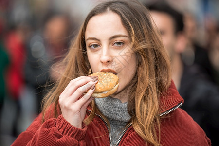 穿着时装服的漂亮可爱女孩在拥挤的街道上行走时吃快餐汉堡包图片