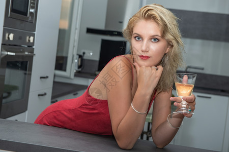 美丽的年轻金发女穿着红裙子在厨房喝白酒图片