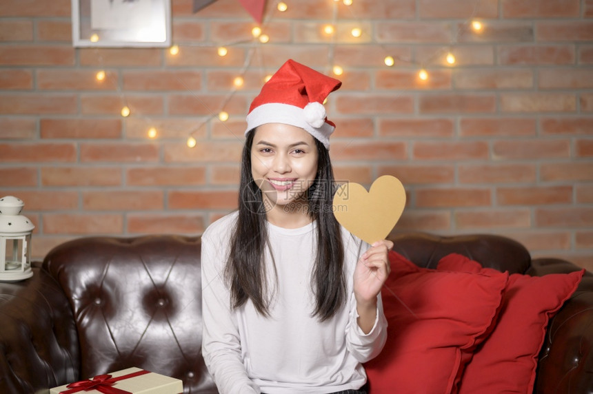 一个戴着红色圣诞老人帽子的微笑年轻妇女展示了圣诞节的心脏形状模型假日概念年轻微笑的女士穿着红色圣诞老人帽子节的心脏形状模型图片