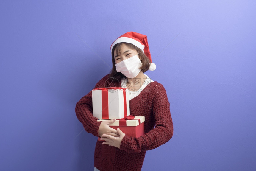 穿着红色圣诞老人帽子戴着外科面具的年轻微笑女人肖像孤立蓝色背景工作室图片