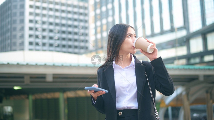 一名身穿黑西装的年轻女商人使用智能手机在城市里端着咖啡图片