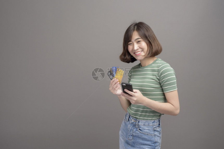 一张持有信用卡和智能手机的年轻美丽亚洲女肖像这些士在灰色背景工作室被孤立图片
