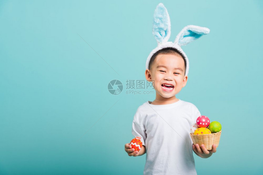 亚洲可爱的小男孩笑着戴兔子耳朵和白色T恤衫站着拿带满复活节蛋的篮子还有一只手拿着带复制空间的蓝背景复活节蛋图片