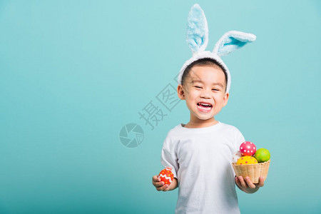 拿对联兔子亚洲可爱的小男孩笑着戴兔子耳朵和白色T恤衫站着拿带满复活节蛋的篮子还有一只手拿着带复制空间的蓝背景复活节蛋背景