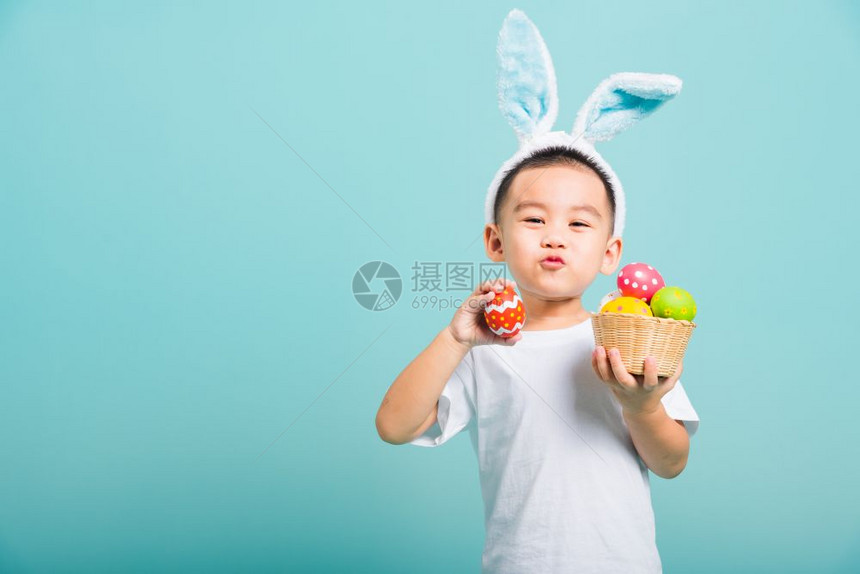 亚洲可爱的小男孩笑着戴兔子耳朵和白色T恤衫站着拿带满复活节蛋的篮子还有一只手拿着带复制空间的蓝背景复活节蛋图片