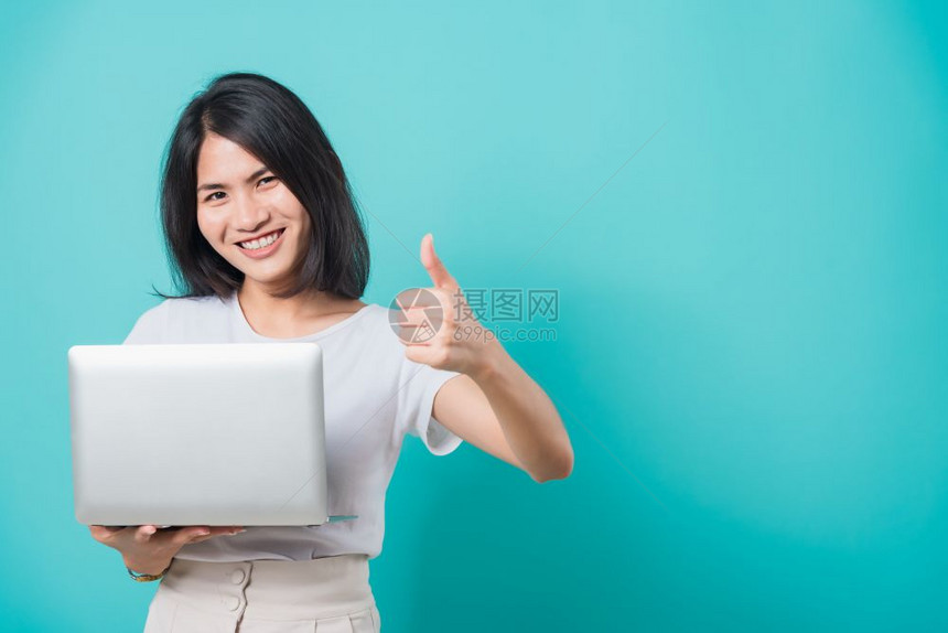 亚洲美丽的年轻女士微笑着白牙站在手持笔记本电脑举起拇指同时在摄影棚拍蓝背景照片并有复制文本的空间图片