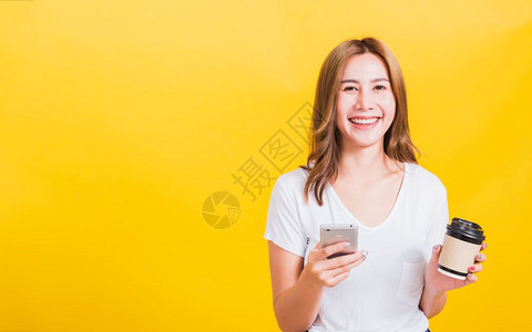肖像亚泰美女快乐的年轻女子站着微笑用手机捧着她的咖啡纸杯望着镜头摄影棚拍摄隔离在黄色背景上有临摹空间背景图片