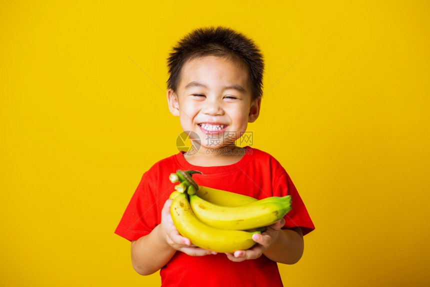 带着红色T恤的笑脸拿着香蕉梳子果拍摄室以黄色背景隔离图片