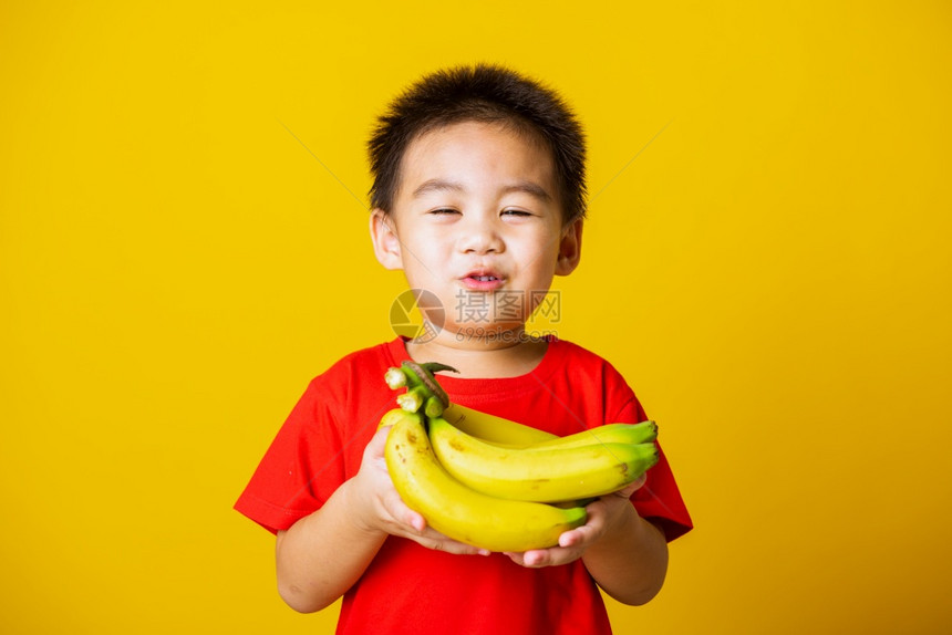 带着红色T恤的笑脸拿着香蕉梳子果拍摄室以黄色背景隔离图片