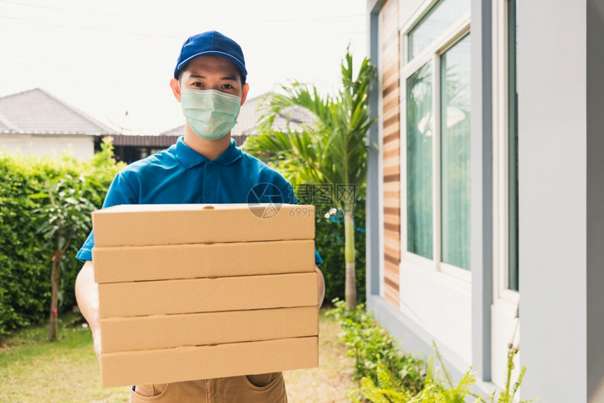 亚洲青年送货员运和持有快餐比萨盒身穿制服在宵禁检疫大流行冠状COVID19下在家庭门口保护面罩服务客户图片