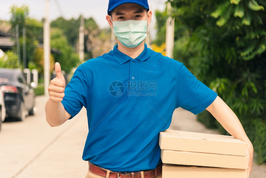 亚洲青年送货员运和持有快餐比萨身穿制服保护面罩戴展示拇指图片