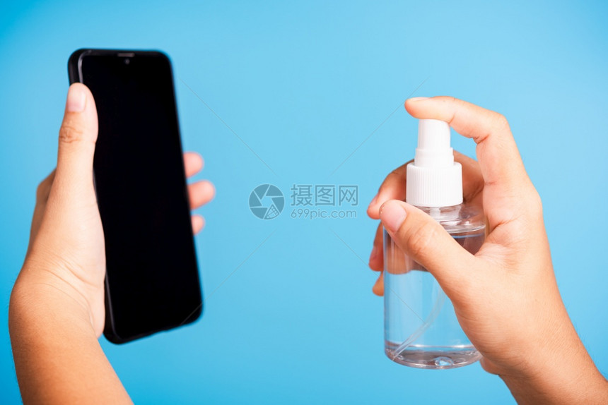 亚洲青年妇女将新闻撒布机净化剂酒精喷洒液射到智能移动电话清洗卫生预防COVID19或冠状保护概念孤立的蓝底图片