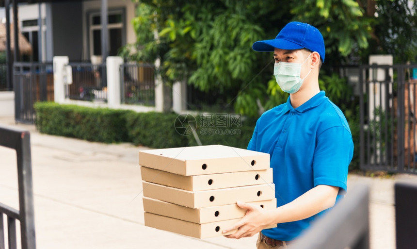 亚洲青年送货员运和持有快餐比萨盒身穿制服在宵禁检疫大流行冠状COVID19下在家庭门口保护面罩服务客户图片