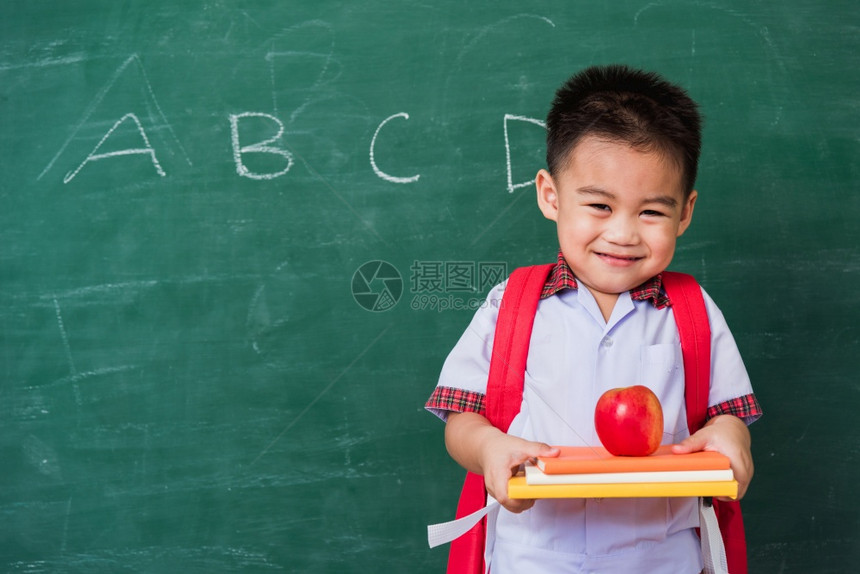 快乐的亚洲可爱小孩男从幼儿园的稚穿着学生制服拿书包红苹果在书上微笑绿色学校黑板上笑第一次到学校教育图片