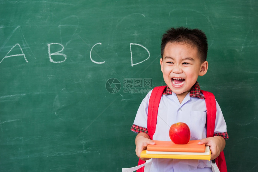 快乐的亚洲可爱小孩男从幼儿园的稚穿着学生制服拿书包红苹果在书上微笑绿色学校黑板上笑第一次到学校教育图片