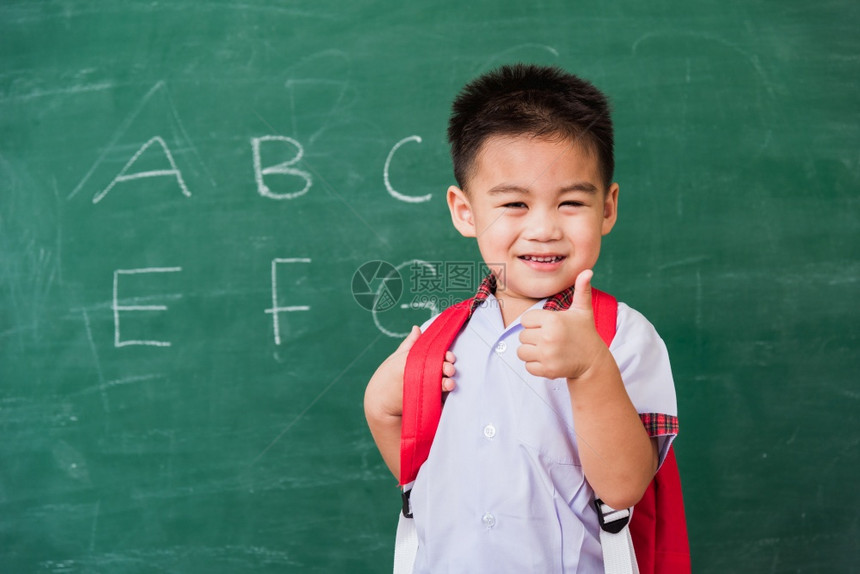 快乐的亚洲可爱小男孩幼儿园穿着校服带微笑的书包在绿色学校黑板上展示手指拇图片