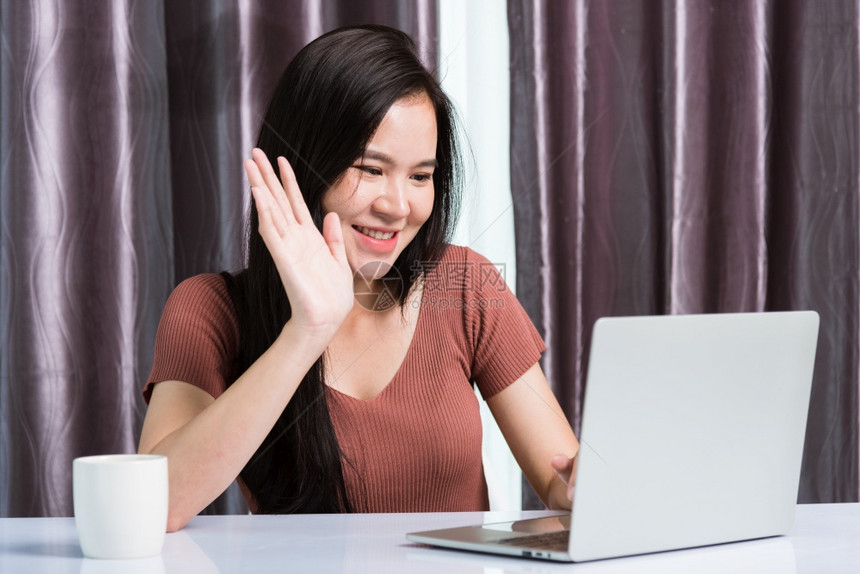 在家工作笑的亚洲快乐商业年轻美丽的女坐在办公桌间视频电话会议上使用网络摄像机手提电脑技术举起手在办公室打招呼图片